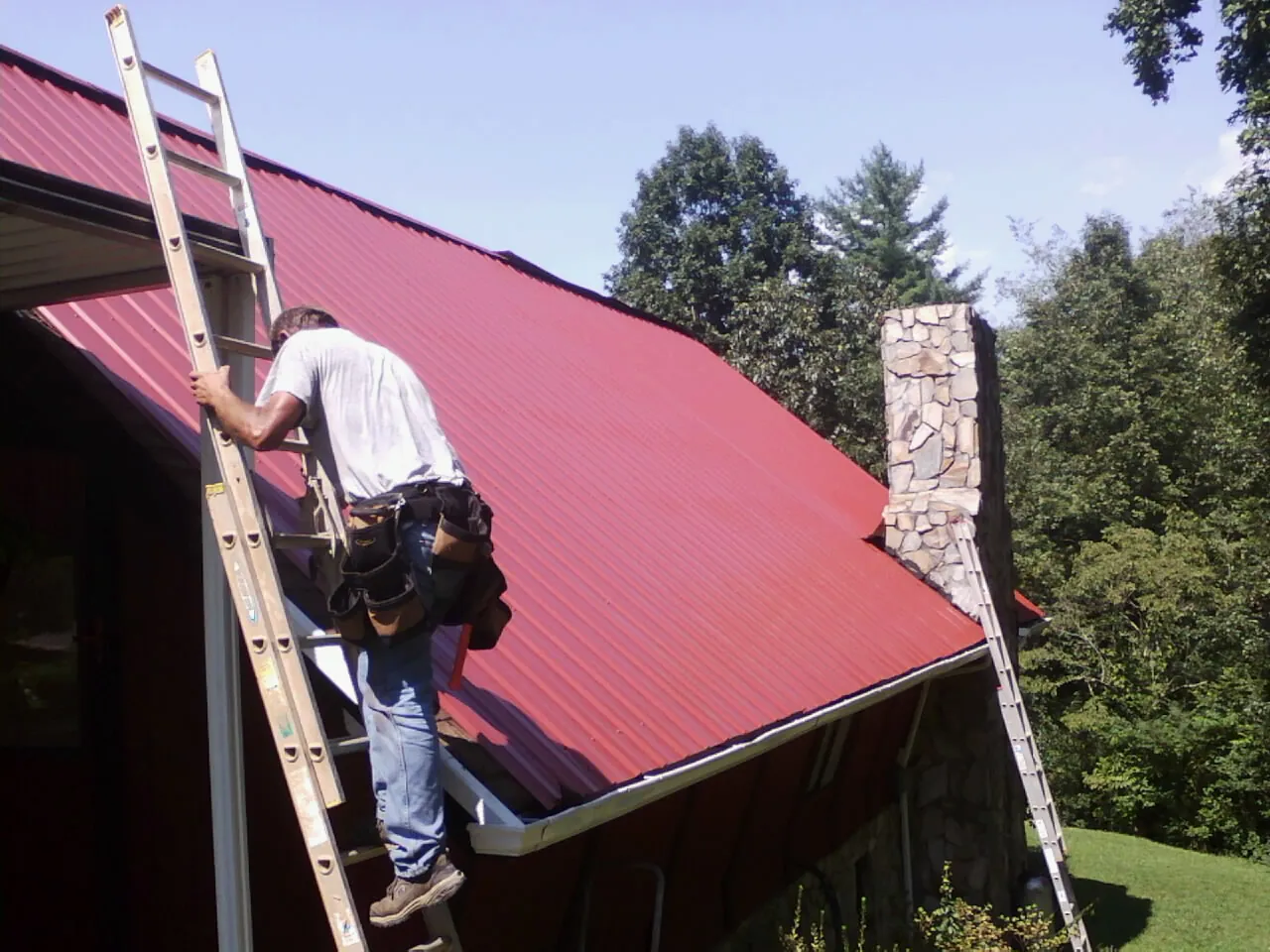 Roofer on a ladder beside a red metal roof and stone chimney — Soffit & Fascia Repair by Roofing Brothers in Oquawka